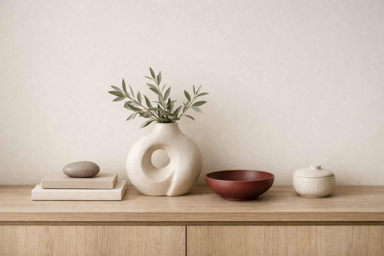  Minimal soft luxury styling vignette with a sculptural ceramic vase, olive branches, neutral books, and natural textures on a light wood console.